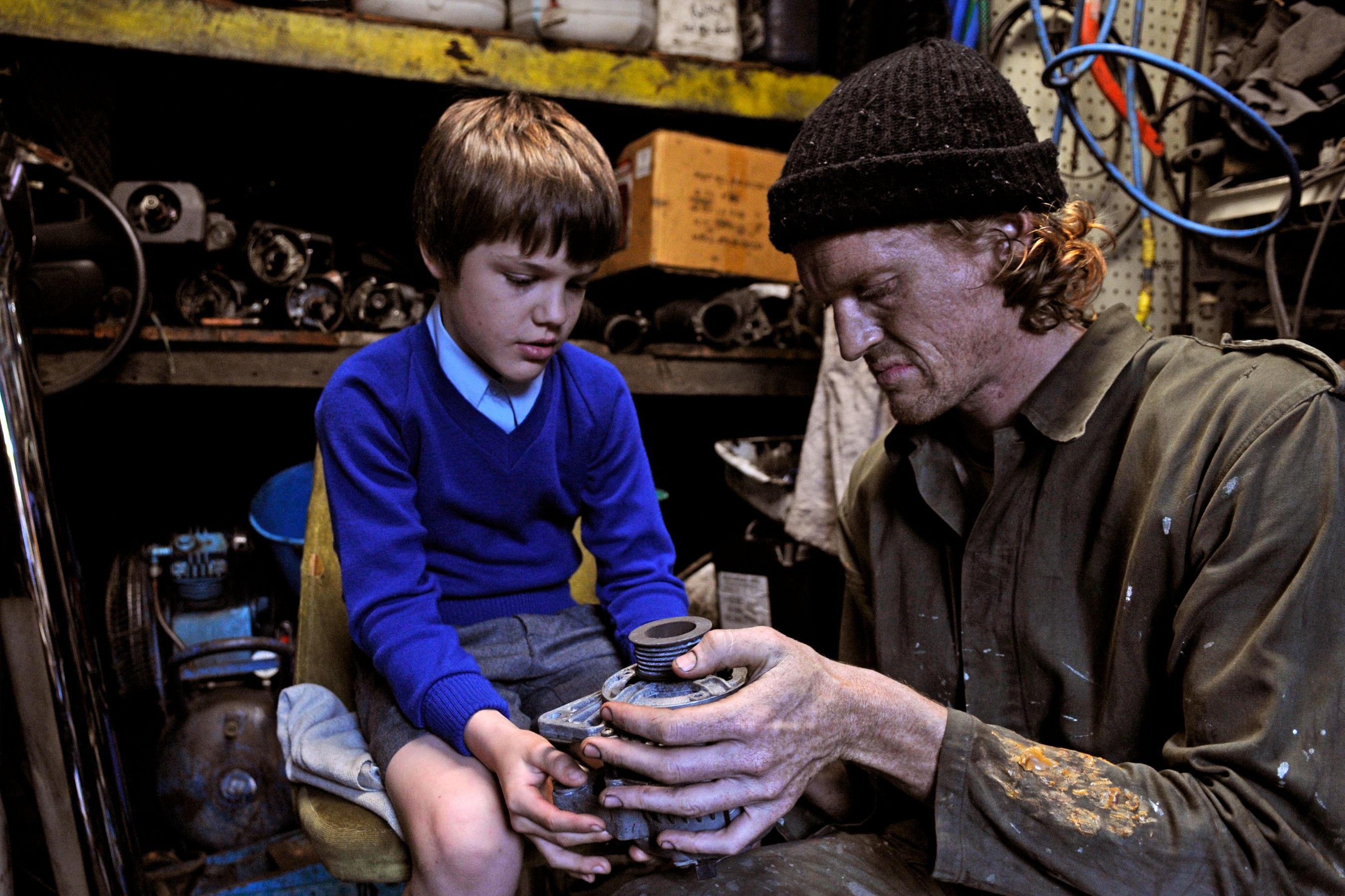 A young boy in a blue sweater and shorts sits on a chair, focused on a mechanical part held by a man dressed in a green jumpsuit and black beanie. The background is cluttered with various tools and parts, indicating a workshop environment. Both appear engaged in a learning moment, with the man sharing knowledge with the boy.