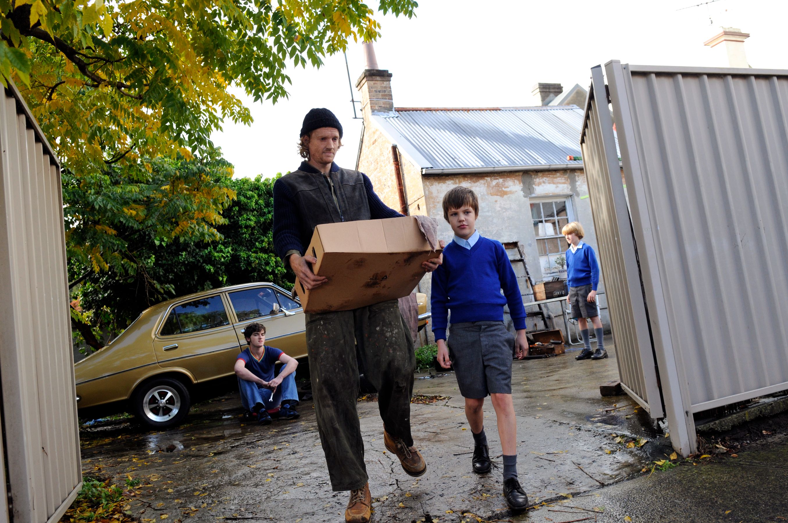 A man carrying a cardboard box walks through a gateway, accompanied by a boy in a blue sweater. In the background, another boy sits on the ground near a vintage car, while a third boy stands beside a building with a weathered facade. The scene is set outdoors, surrounded by greenery and scattered leaves.