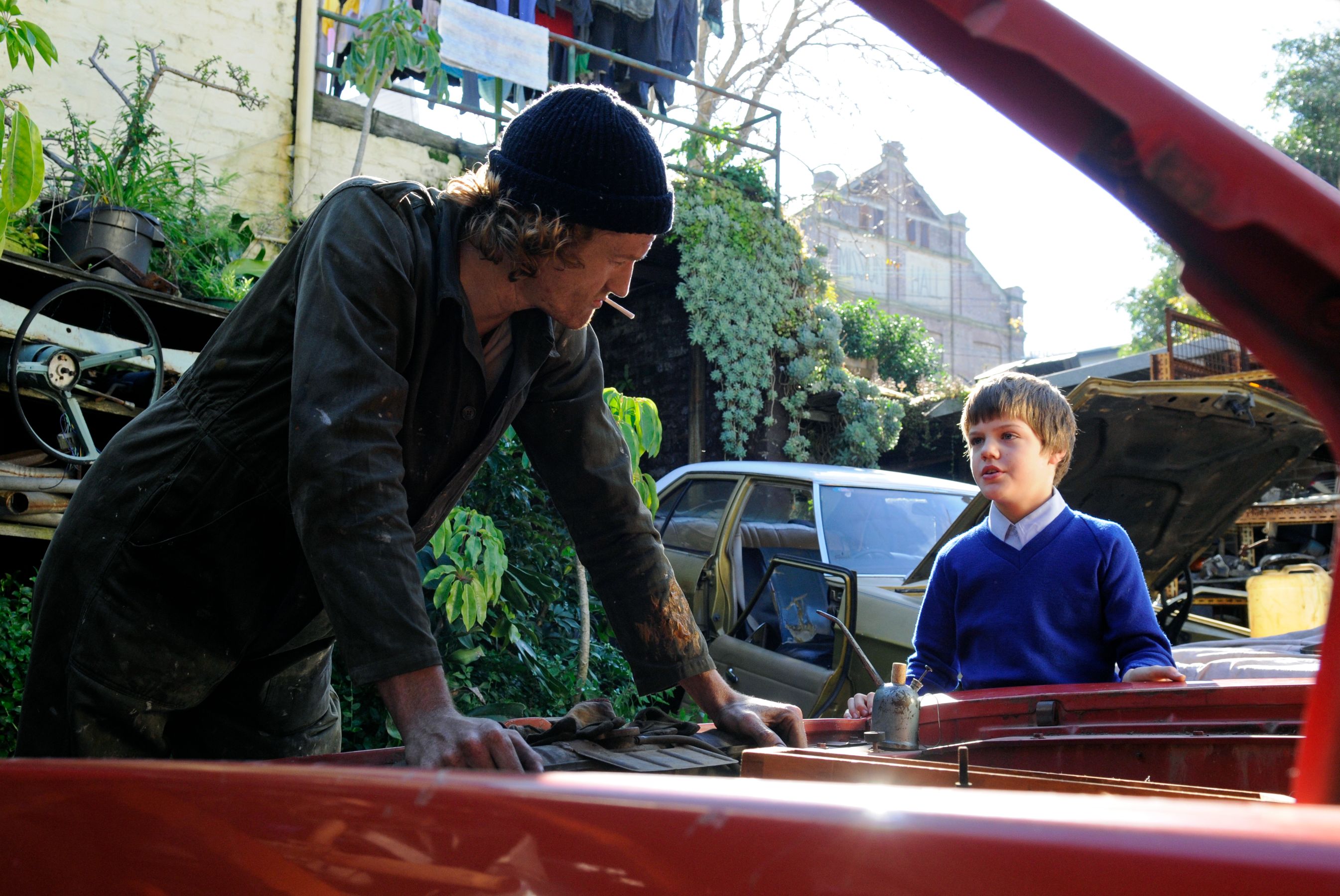 A man in a dark, dirty outfit leans over a car engine, speaking intently to a young boy wearing a blue sweater and white shirt. The background features overgrown plants and cluttered cars, creating an atmosphere of a makeshift workshop.
