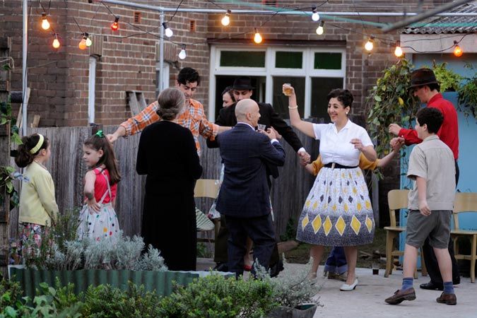 A lively outdoor gathering shows a group of people dancing and enjoying each other's company under string lights. The scene includes adults and children, with some dressed in vintage clothing, creating a cheerful and nostalgic atmosphere.