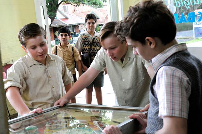 Three boys are gathered around a pinball machine, intently focused on the game. A fourth boy stands in the background, watching them with a curious expression. The setting appears to be a vintage arcade or game room, with visible greenery outside.