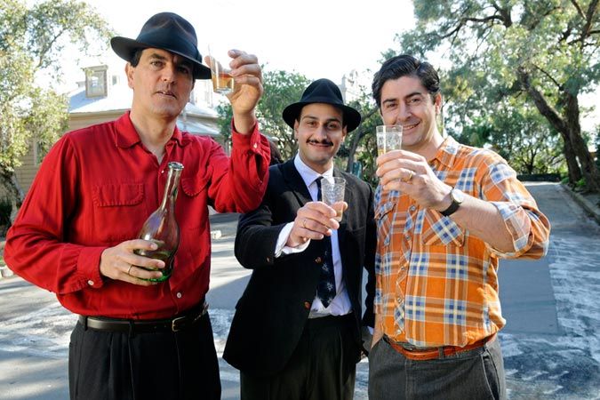 Three men stand outdoors, each holding drinks. One in a red shirt and black hat holds a bottle, the second in a black suit with a tie has a glass, and the third in an orange plaid shirt also raises a glass. They appear to be celebrating and smiling, with trees and buildings in the background.