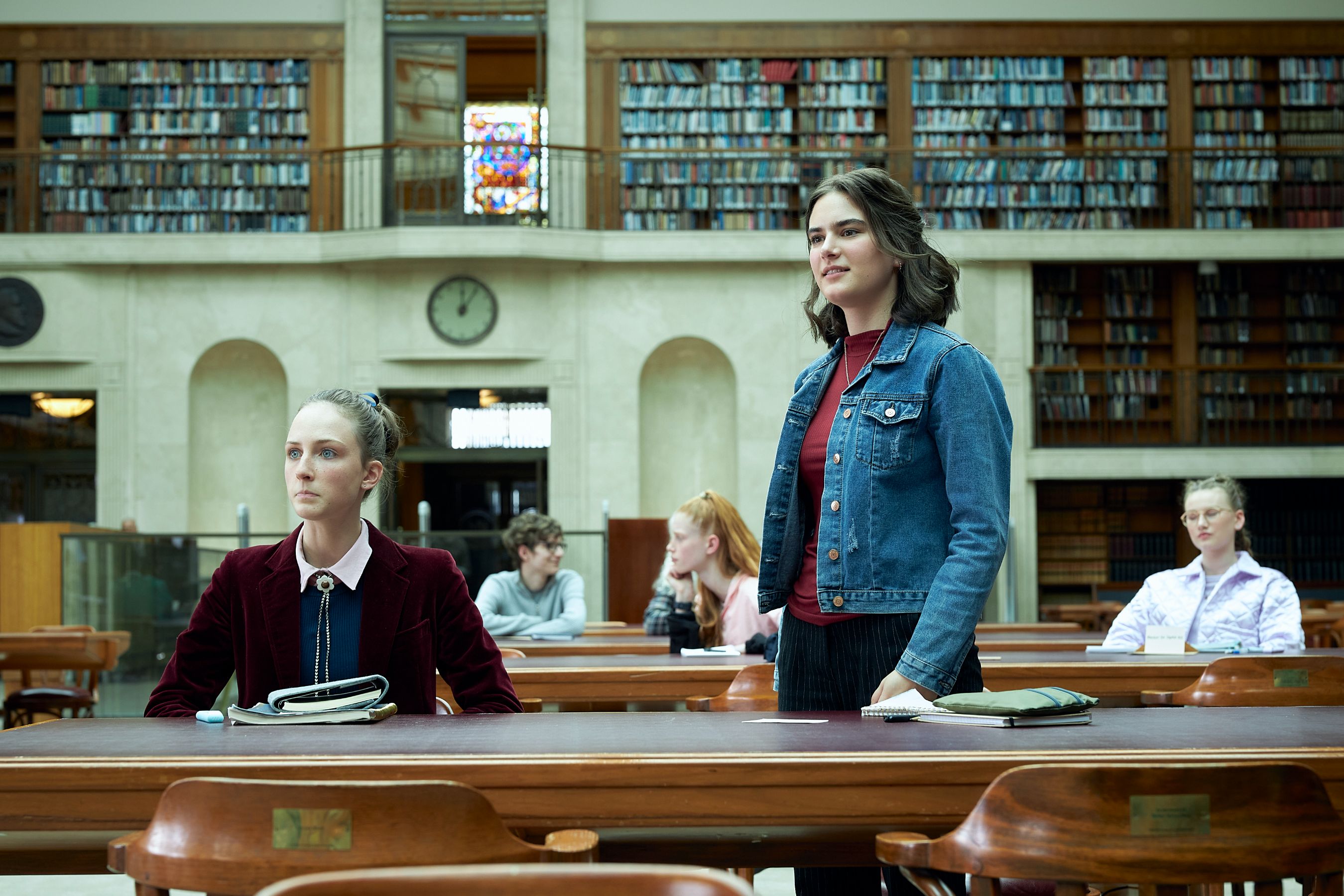 THE PM'S DAUGHTER_Sadie (Natalie English)_Cat (Cassandra Helmot)_Ep3_SD33_0145 Two young women sit in a library surrounded by wooden tables and shelves filled with books. One woman sits with a focused expression, while the other stands, looking confidently forward. Other students are scattered around, engaged in study. The atmosphere is quiet and studious, highlighted by a large clock on the wall and colorful stained glass windows above.