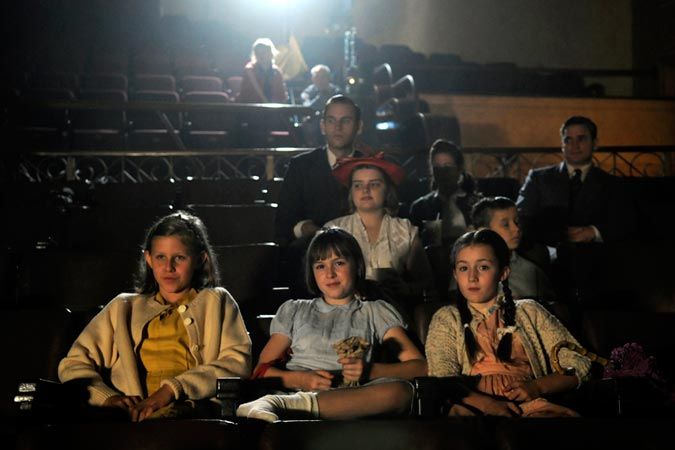 A group of children sits in a dimly lit theater, their expressions a mix of curiosity and anticipation. The older girls are dressed in 1940s attire, while adults are visible in the background, also focused on the performance.