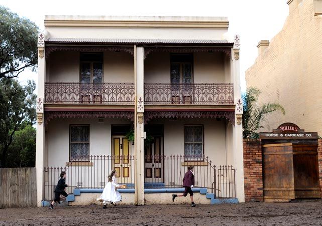 Two children run through a muddy area in front of a two-story Victorian-style building with decorative ironwork on the balconies. One child is dressed in white, while the other wears dark clothing; a sign for "Miller's Horse & Carriage Co." can be seen on the right side of the image.