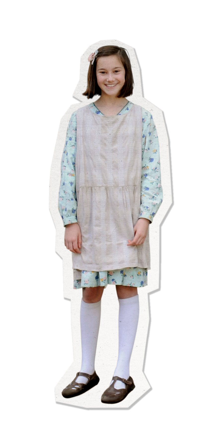 A young girl stands smiling, wearing a floral-patterned dress layered with a beige apron. She has short brown hair adorned with a flower clip and is dressed in knee-high white socks and brown shoes.