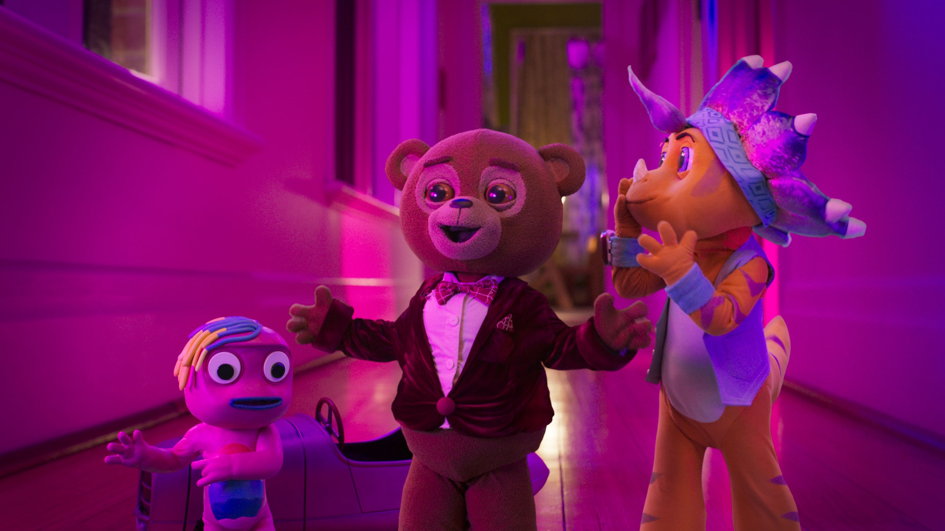 Three colorful animated characters stand in a brightly lit hallway with purple hues. A bear in a red tuxedo smiles widely, while a small blue figure with colorful hair displays a surprised expression, and a dinosaur-like creature leans in playfully.
