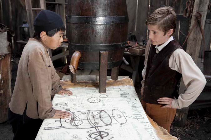 Two young boys are focused on a large wooden table covered with sketches and notes, discussing what appears to be an invention. Behind them stands a large wooden barrel with a spout, and the setting suggests a workshop filled with tools and materials.