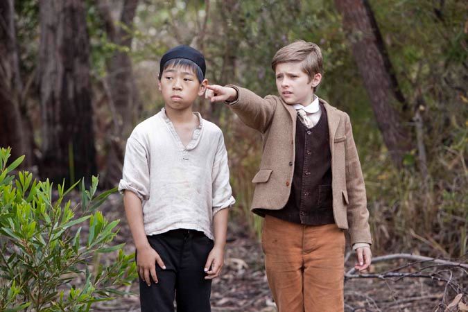 Two young boys stand in a wooded area. One boy, wearing a light-colored shirt and dark pants, appears serious while the other boy, dressed in a brown suit and cream-colored shirt, points at something with a concerned expression. Surrounding them are trees and shrubs.