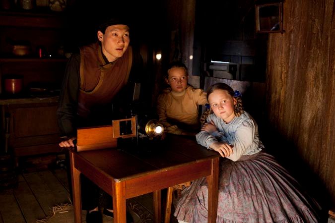Three children and a young man gather around a small table with an antique projector. The room is dimly lit, creating a warm, nostalgic atmosphere as they intently watch the light projected from the device. One girl has curly hair and wears a blue blouse, while another girl, sitting next to her, appears focused. The young man, dressed in dark clothing, leans slightly toward the projection, sharing the moment with the group.