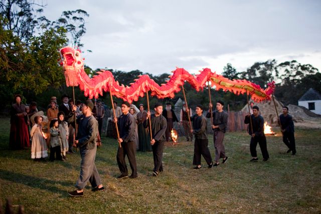 A group of performers dressed in dark clothing holds a long, vibrant red and gold dragon costume, parading through a grassy area at dusk. In the background, onlookers watch, some wearing historical attire, as the scene is illuminated by torchlight.