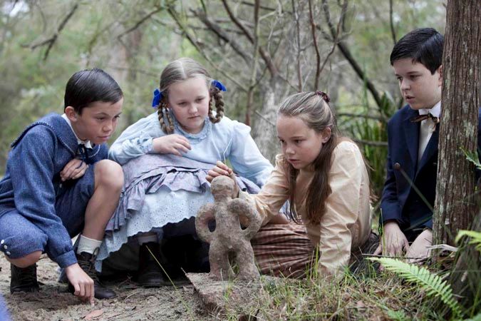Four children are gathered around a small stone sculpture of a figure in a wooded area. They are intently studying the sculpture, with expressions of curiosity and intrigue; two girls wear dresses, while the boys are in suits.