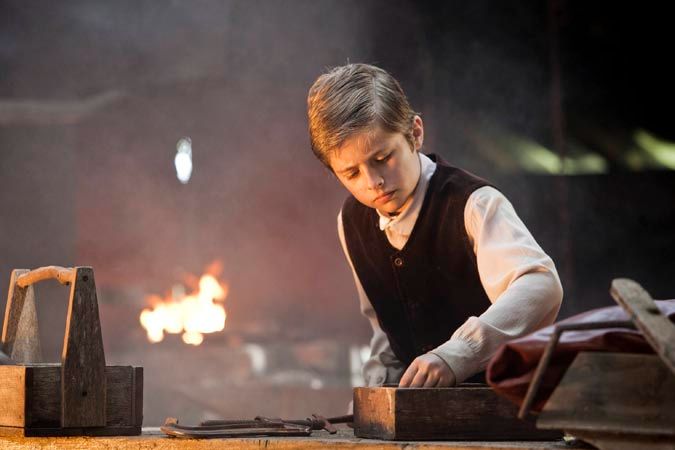 A young boy, dressed in period clothing, concentrates on a wooden workbench in a dimly lit workshop filled with smoke. The backdrop features a warm glow from a fire, suggesting a lively but gritty environment.