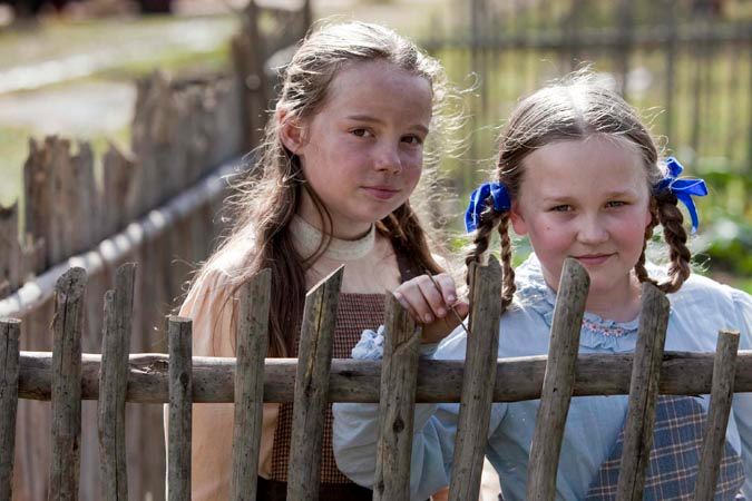 Two young girls stand together near a rustic wooden fence, smiling warmly. One girl has long, wavy hair and wears a brown dress, while the other has hair tied in pigtails adorned with blue ribbons, dressed in a light blue outfit.