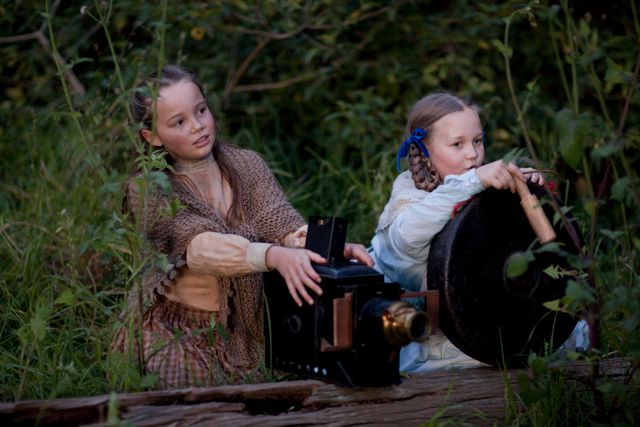 Two young girls are sitting in a forest, appearing engaged in a playful scene. One girl, wearing a brown scarf and a beige shirt, operates an old-fashioned camera, while the other girl, dressed in white and decorated with blue ribbons in her hair, holds a large circular object, possibly a lens or a reflector. Surrounding them are lush green plants, adding to the enchanting atmosphere.
