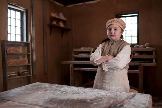 A young child stands confidently with arms crossed in a rustic bakery, wearing a beige baker's hat and simple clothing. Shelves in the background hold various types of bread, and flour dust is visible on the wooden table in front of them.