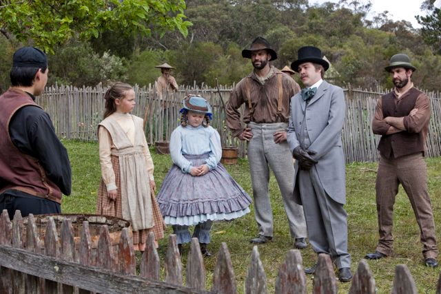 A group of six people stands in a rural setting near a wooden fence. The group includes two girls in modest, vintage dresses, one wearing a hat, and four adults dressed in historical clothing, including a man in a suit and a top hat, and another in work clothes. They are gathered around a wooden barrel, with a natural landscape visible in the background.