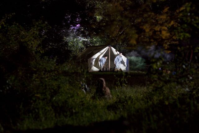 A tent is set up in a dimly lit area surrounded by tall grass and trees, with faint shadows of figures visible near the entrance. Ambient light from an unseen source casts an eerie glow around the scene, suggesting a nighttime setting.