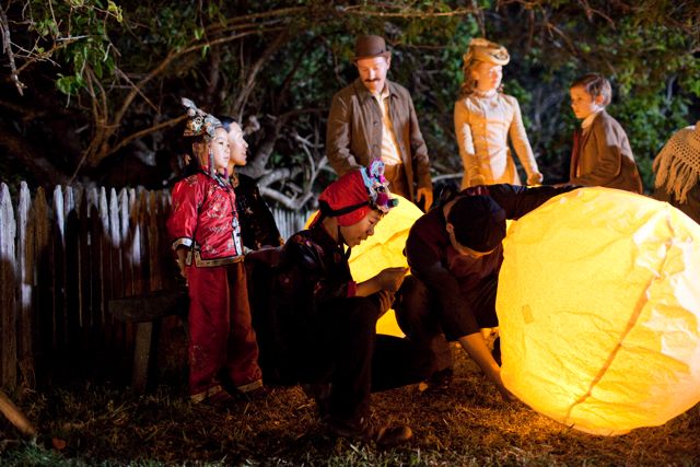 A group of children and adults gather outdoors at night, illuminated by the warm glow of a large lantern being prepared by two individuals. Some children are dressed in colorful costumes, while others observe with curious expressions, surrounded by trees and a rustic fence.