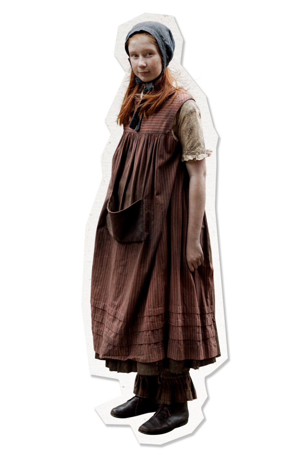 A young girl with long red hair stands wearing a traditional, long brown dress with vertical stripes and a pocket. She is also wearing a lace-trimmed blouse and dark shoes, with a dark scarf tied around her head and a necktie. The backdrop is plain, emphasizing her vintage attire.