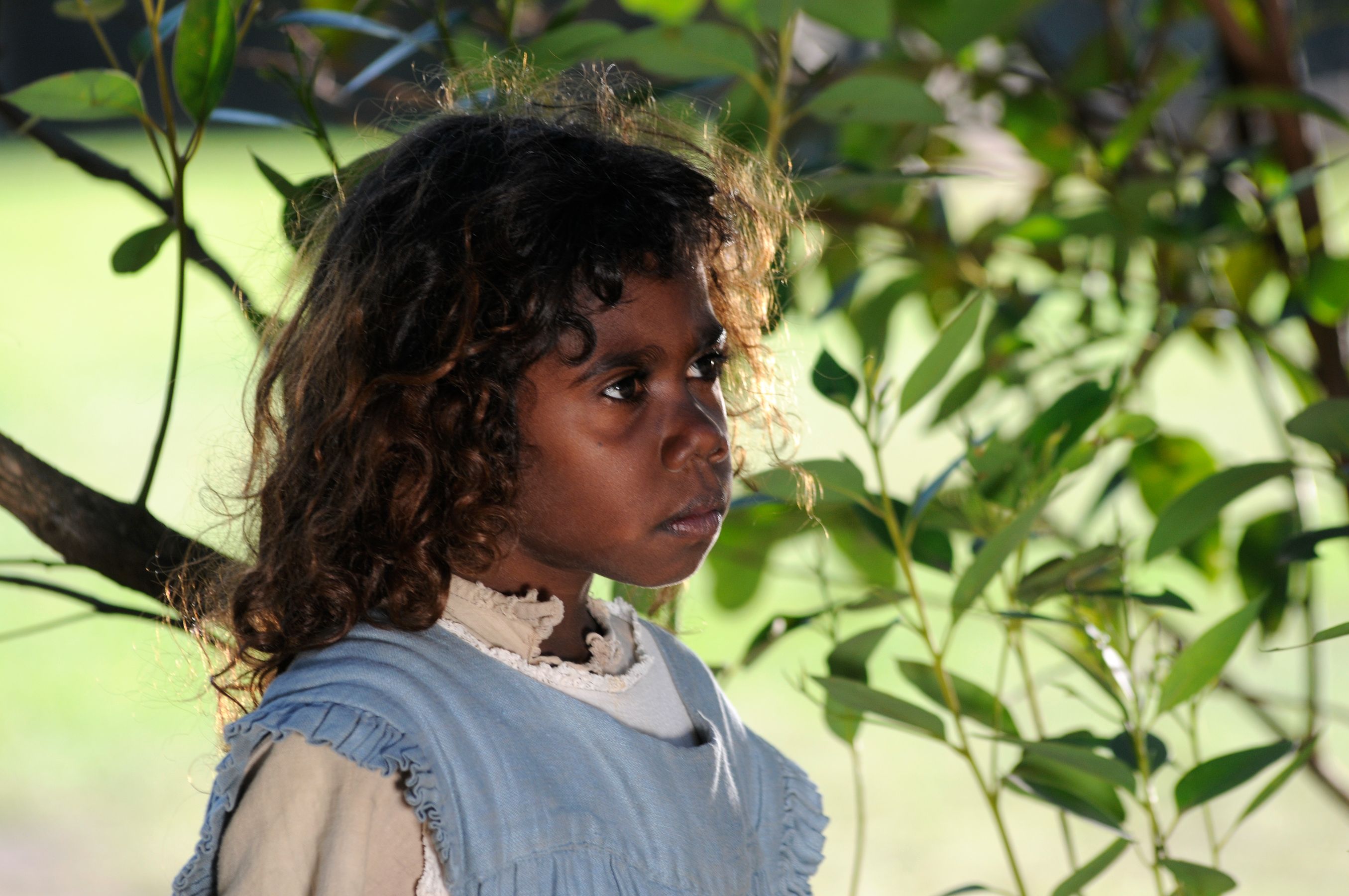 A girl with curly hair looks intently while standing near lush green foliage. She wears a light blue dress and has a serious expression on her face.