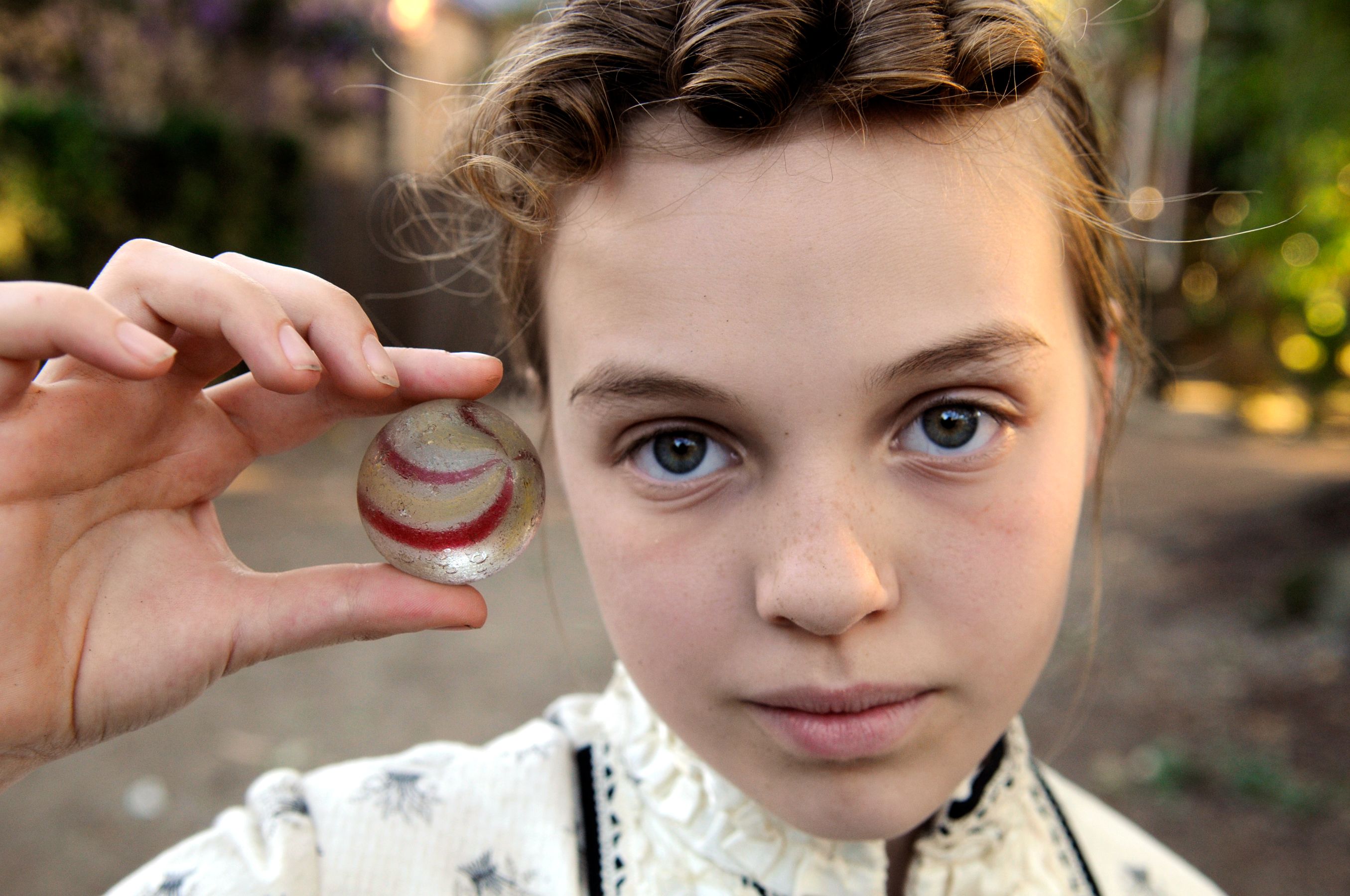 A young girl with curly hair holds a glass marble with red swirls against a blurred outdoor background. She gazes directly at the camera with striking blue eyes and a serious expression, wearing a patterned shirt.