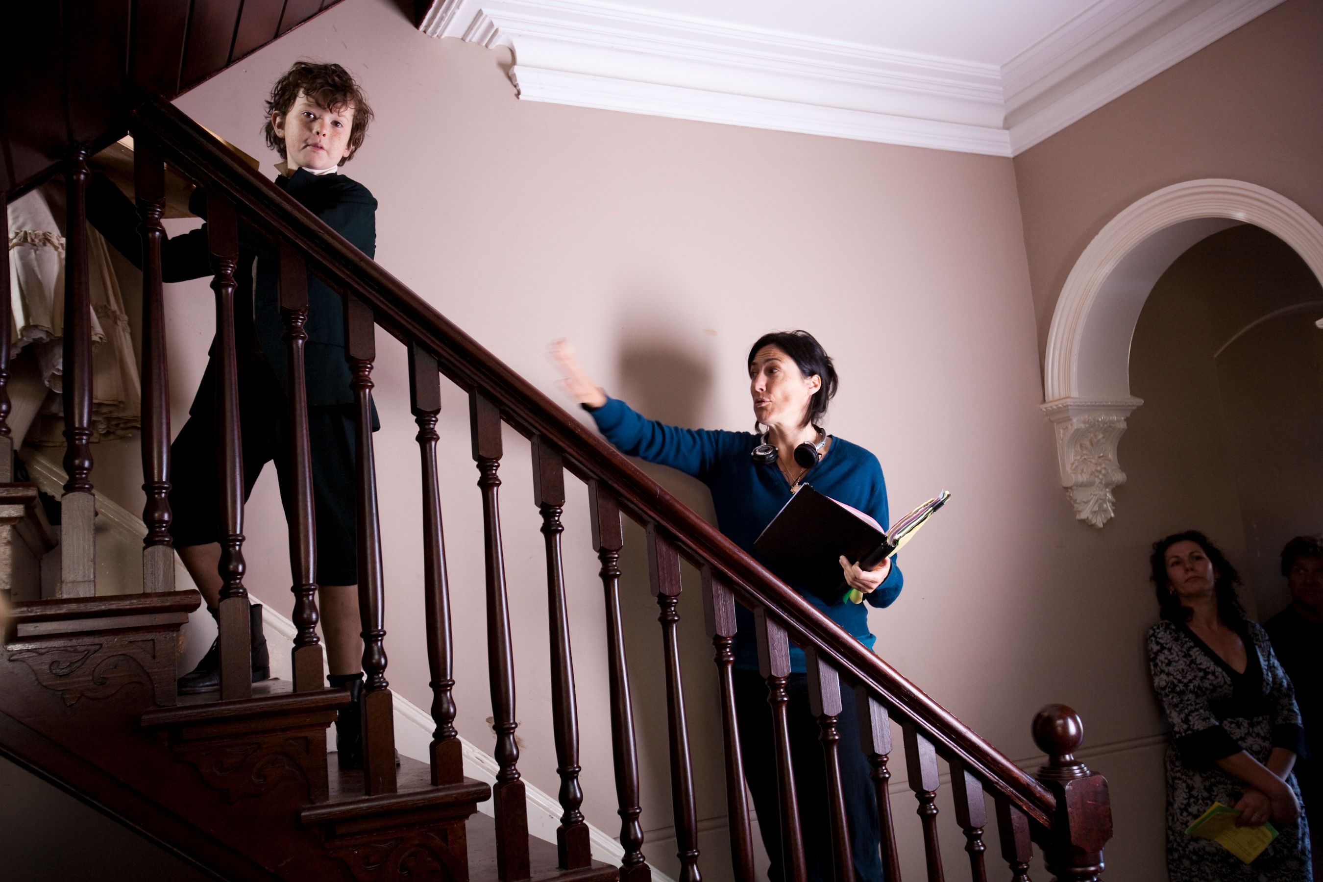 A young boy in a black outfit stands on the stairs, looking down at an adult woman at the bottom, who gestures while holding a binder. Behind her, two other individuals observe the interaction, creating a scene that appears to involve direction or discussion. The setting has warm tones and classic architectural details.