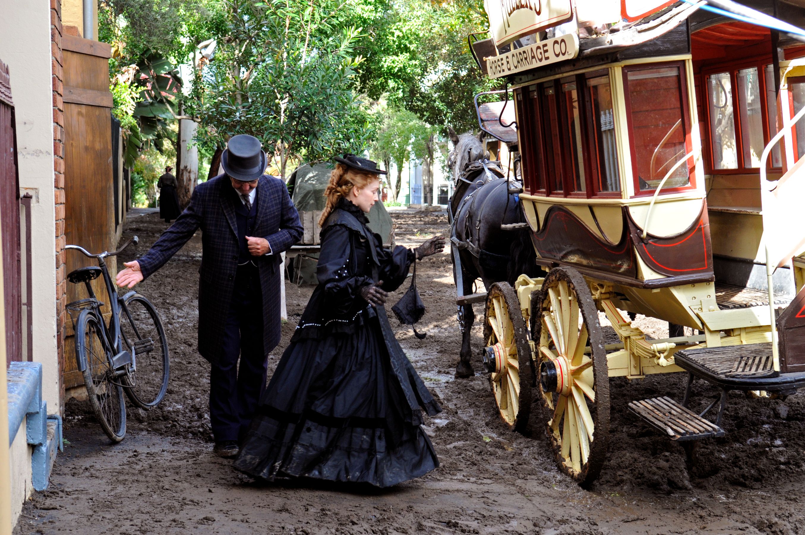 A man in a formal suit and top hat gestures toward a vintage bicycle while a woman in a long, elegant black dress and hat approaches a horse-drawn carriage. The scene is set in a muddy lane surrounded by trees and buildings typical of a historical era.