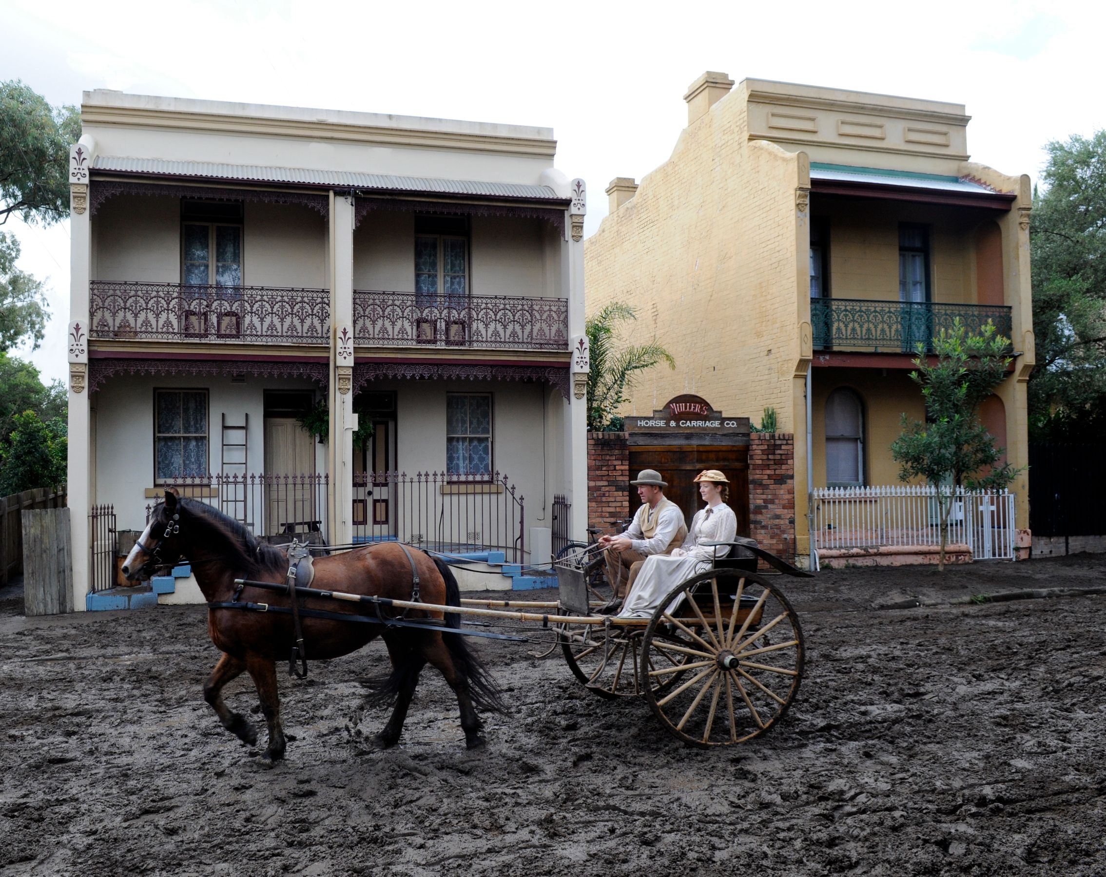 A horse-drawn carriage is seen moving through a muddy area in front of two historic buildings, featuring ornate balcony designs and a sign for a horse and carriage company. Two individuals dressed in period clothing sit in the carriage, evoking a sense of the past.
