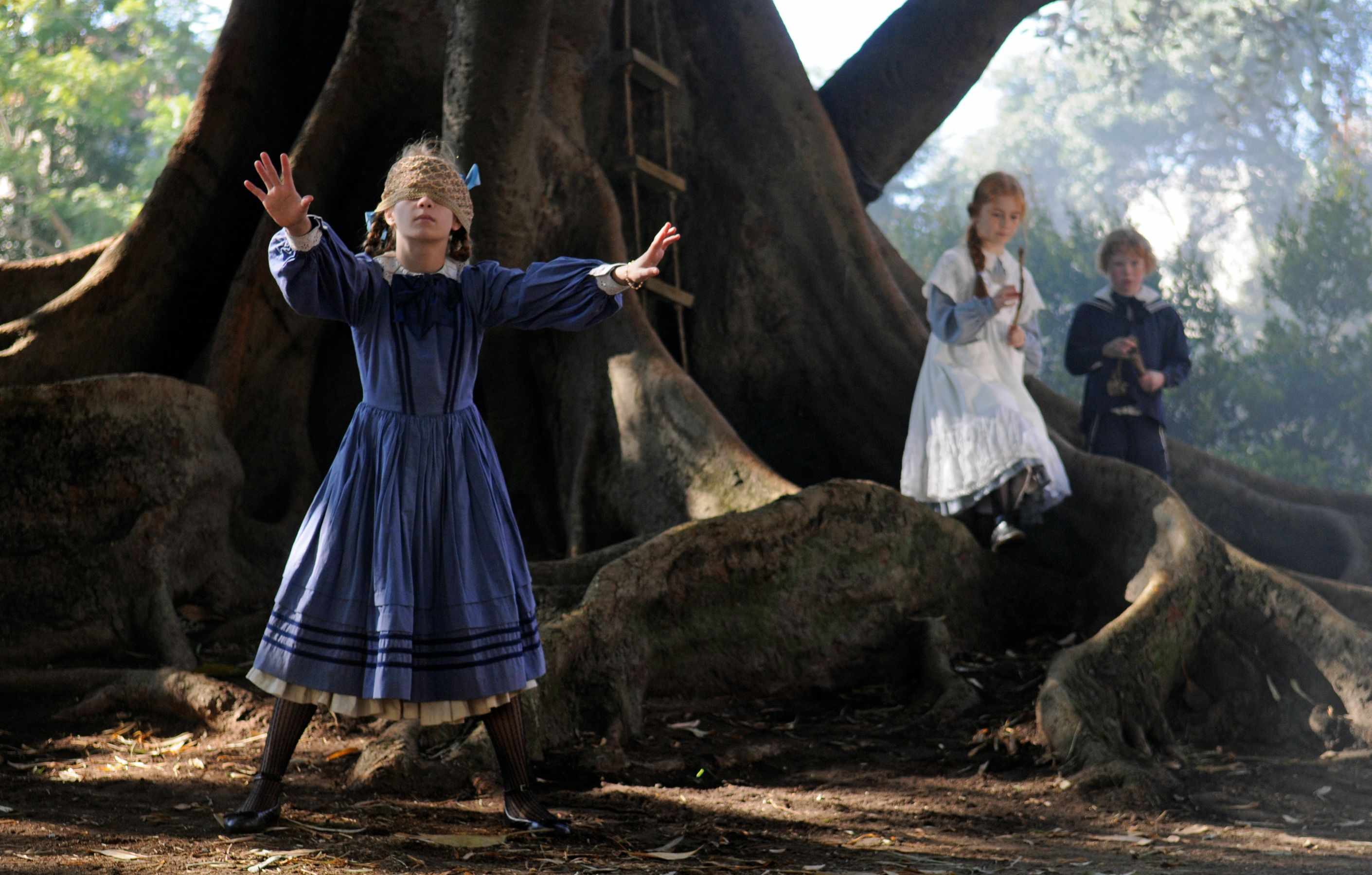 A girl in a blue dress stands with her arms outstretched, wearing a blindfold beneath a large tree, while two other children watch nearby. One child is dressed in white and holding a stick, and the other is in navy, both standing on the ground amid roots and foliage.