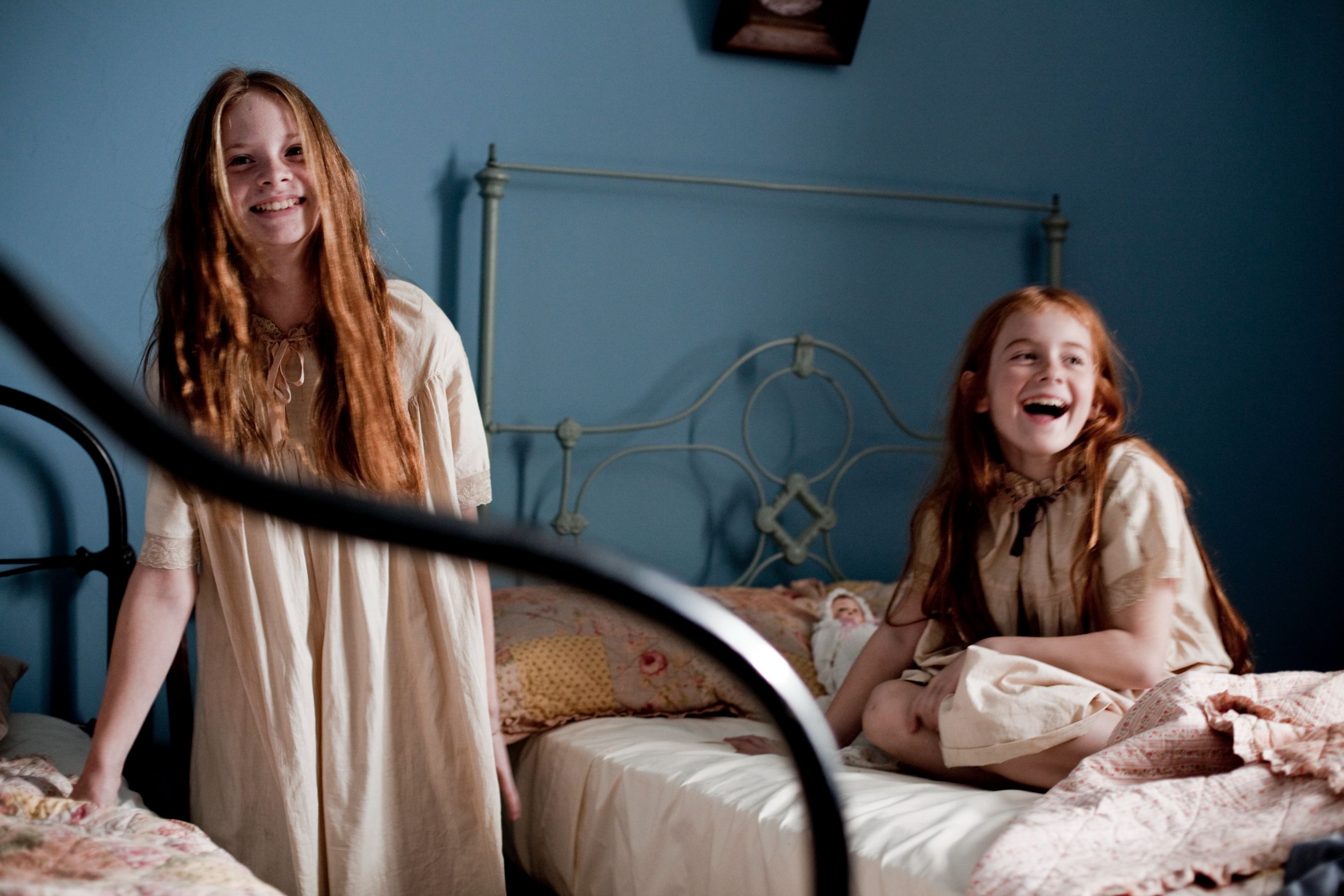 Two young girls with long hair, dressed in light-colored nightgowns, are in a softly lit bedroom with blue walls. One girl is standing and smiling, while the other is sitting on a bed, laughing joyfully. The room has two beds, and the atmosphere appears cheerful and playful.