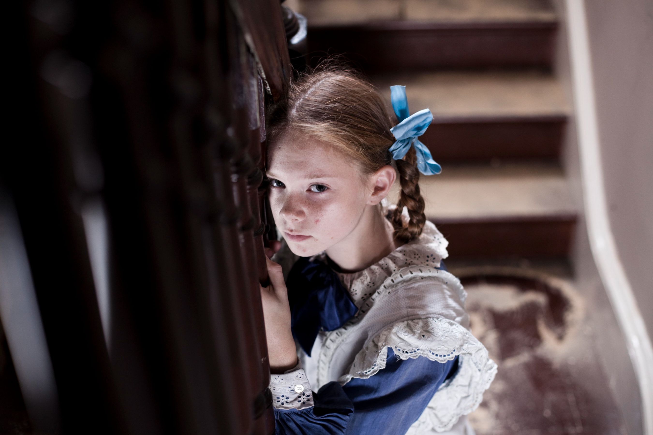 A young girl with braided hair and a blue ribbon gazes intently at the viewer while holding onto a wooden railing. She wears a vintage-style dress with lace detailing, and the setting features a staircase with worn steps, conveying a sense of intrigue or unease.