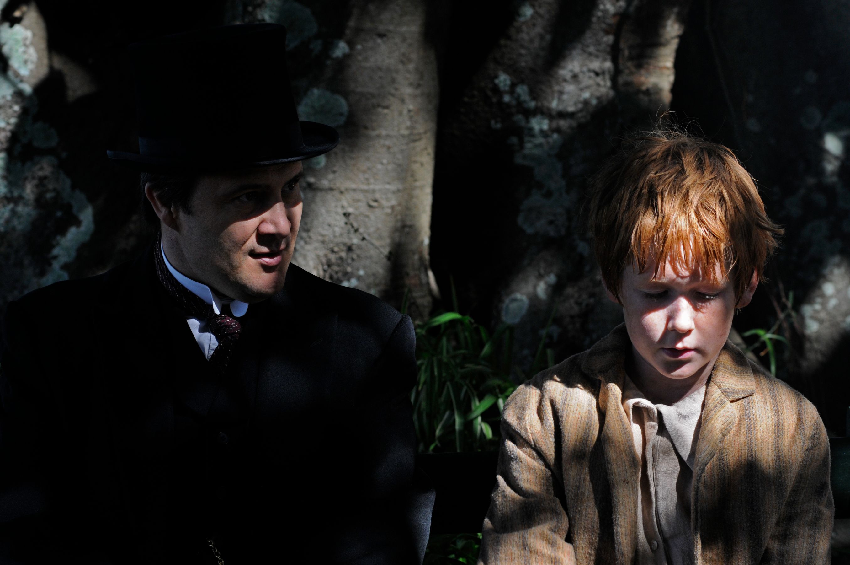 A well-dressed man in a black top hat and dark coat is seated beside a young boy with short red hair, who is wearing a light-colored shirt and a striped brown jacket. The scene is shaded by a large tree, creating a contrast between light and shadow, as the two appear deep in thought or conversation.