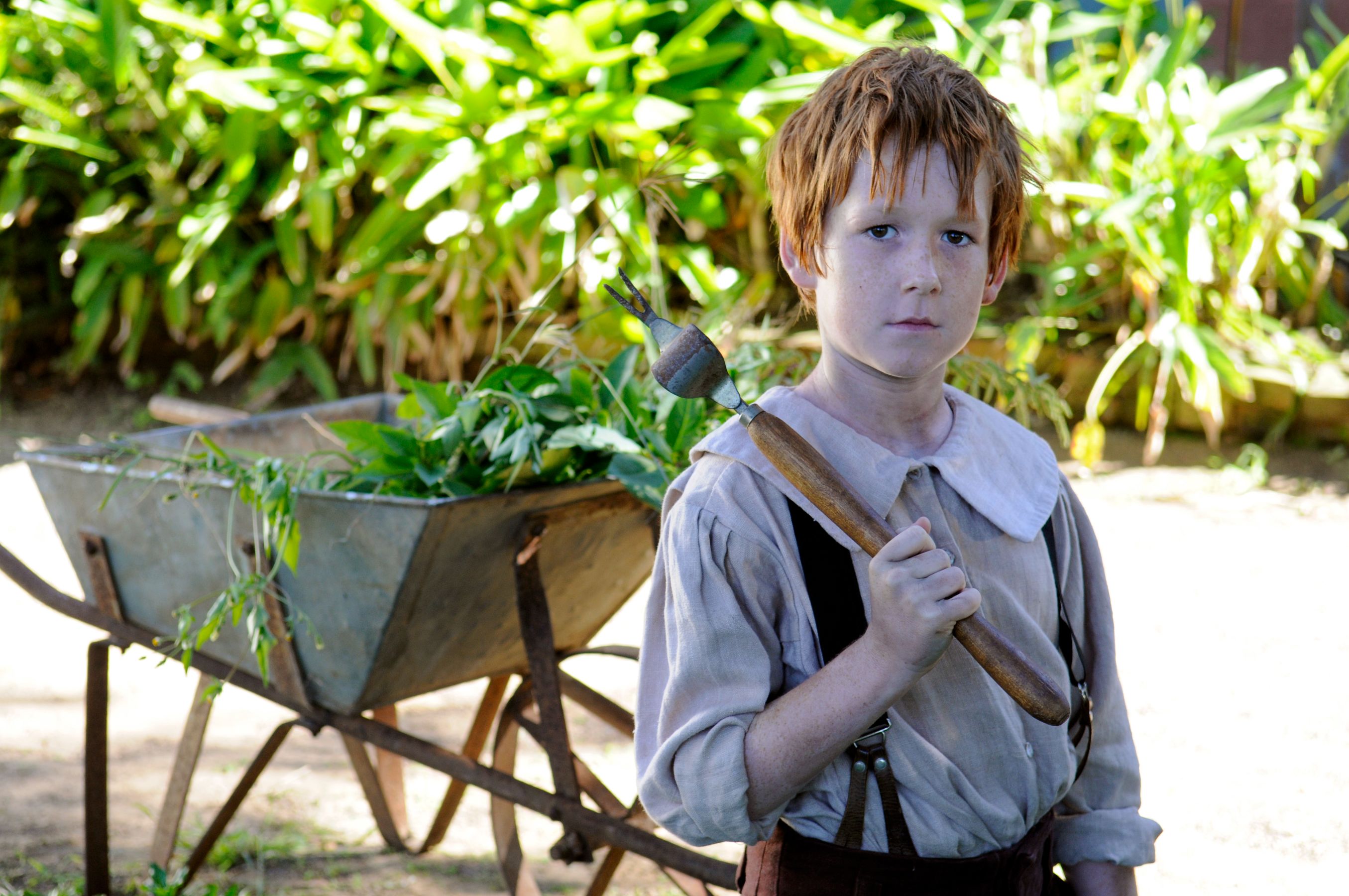 A young boy with red hair stands holding a small gardening tool, wearing a simple shirt with a collar and suspenders. Behind him is a wheelbarrow filled with greenery, set against a backdrop of lush plants.
