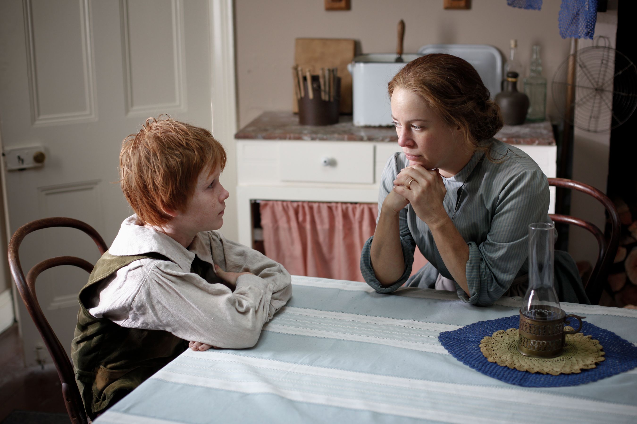 A young child with short, messy red hair sits at a table, looking intently at a woman in period clothing who appears to be deep in thought. The kitchen setting features a vintage decor with a white cabinet, a cloth draped over an opening, and a small oil lamp on a blue crocheted mat.