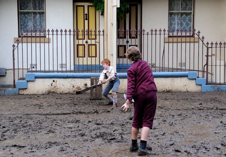 Two boys play in a muddy yard, with one boy holding a shovel and appearing to dig, while the other stands nearby. The background features a house with a railing, and the scene conveys a sense of childhood playfulness, despite the muddy surroundings.
