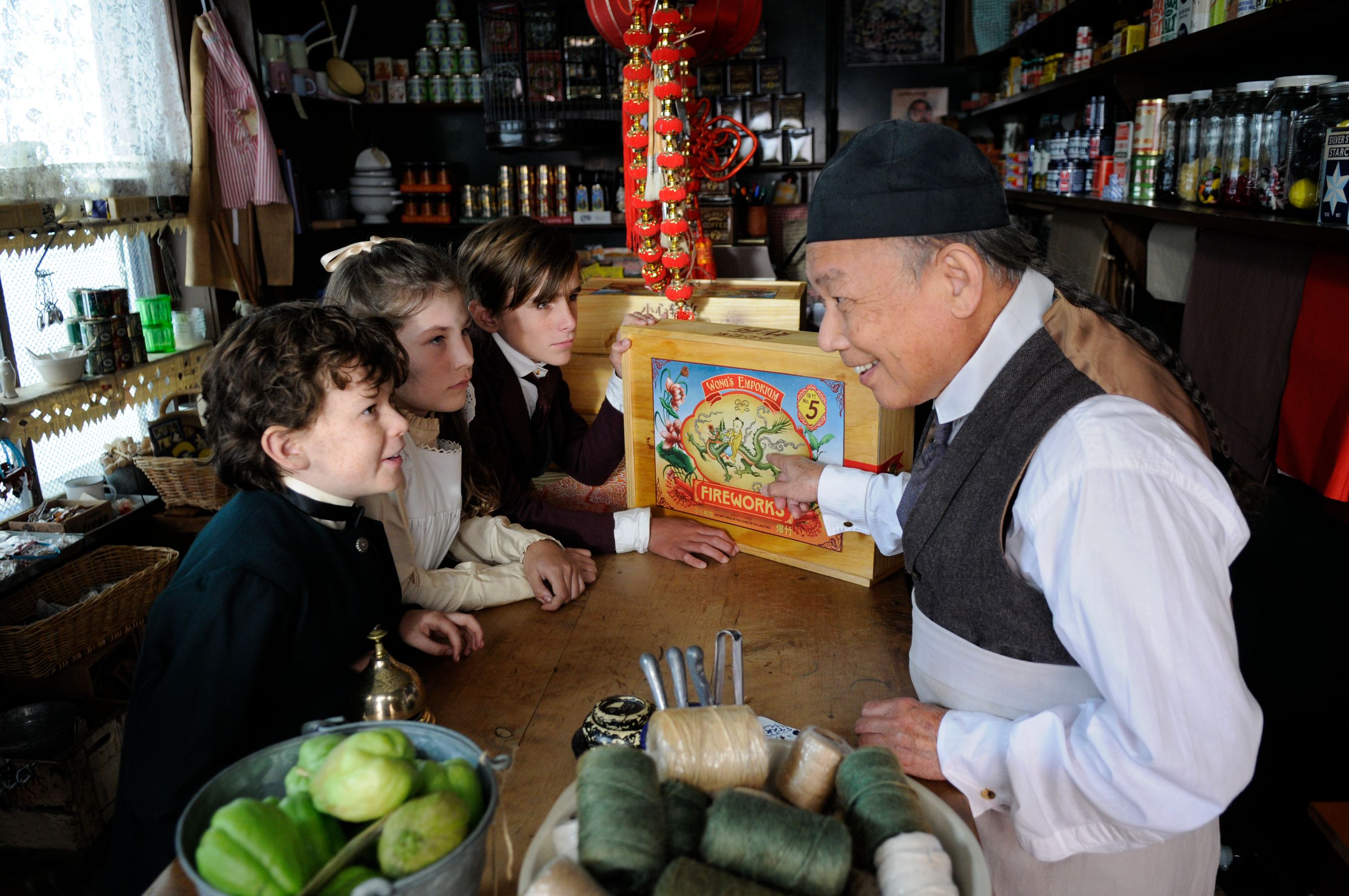 A shopkeeper gestures towards a colorful box labeled "Fireworks" while interacting with three curious children at a wooden counter. The store's shelves are filled with various goods, and old-fashioned items create a charming atmosphere.