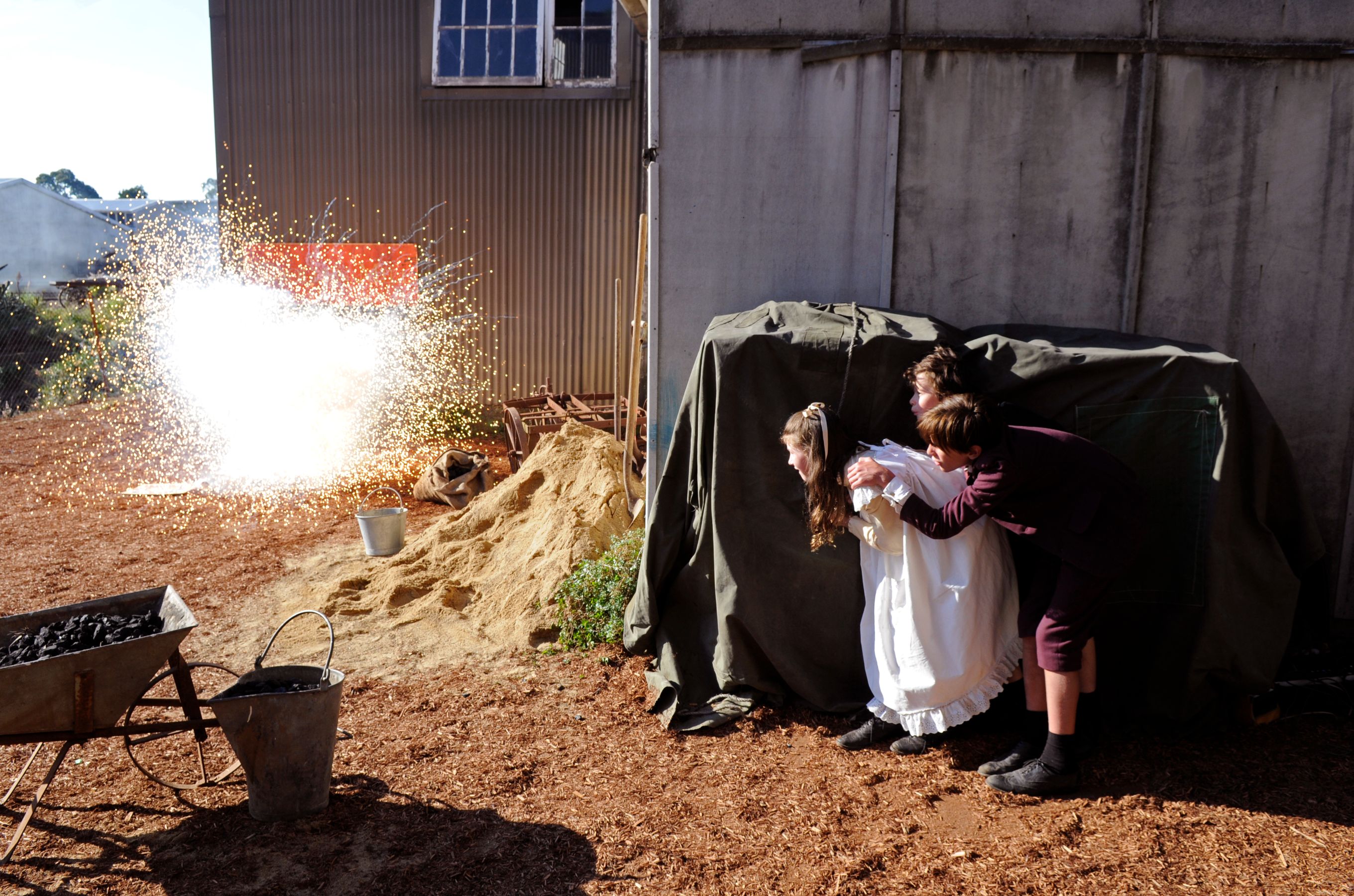 Two children peek out from behind a green tarp, their expressions a mix of curiosity and apprehension. A bright explosion occurs nearby, sending sparks and light into the air against the backdrop of a rustic building and piles of dirt.