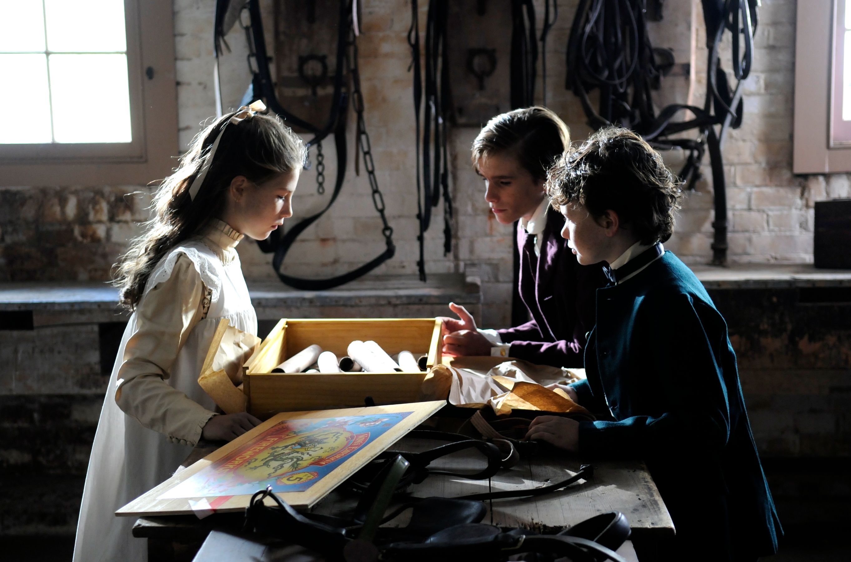 Three children are gathered around a wooden table in a dimly lit room, examining a box filled with rolled papers. One girl, in a light dress and with long hair, focuses intently on the contents while the two boys, dressed in dark clothing, look on with curiosity. Various objects and tools are visible on the table, adding to the historical ambiance of the scene.
