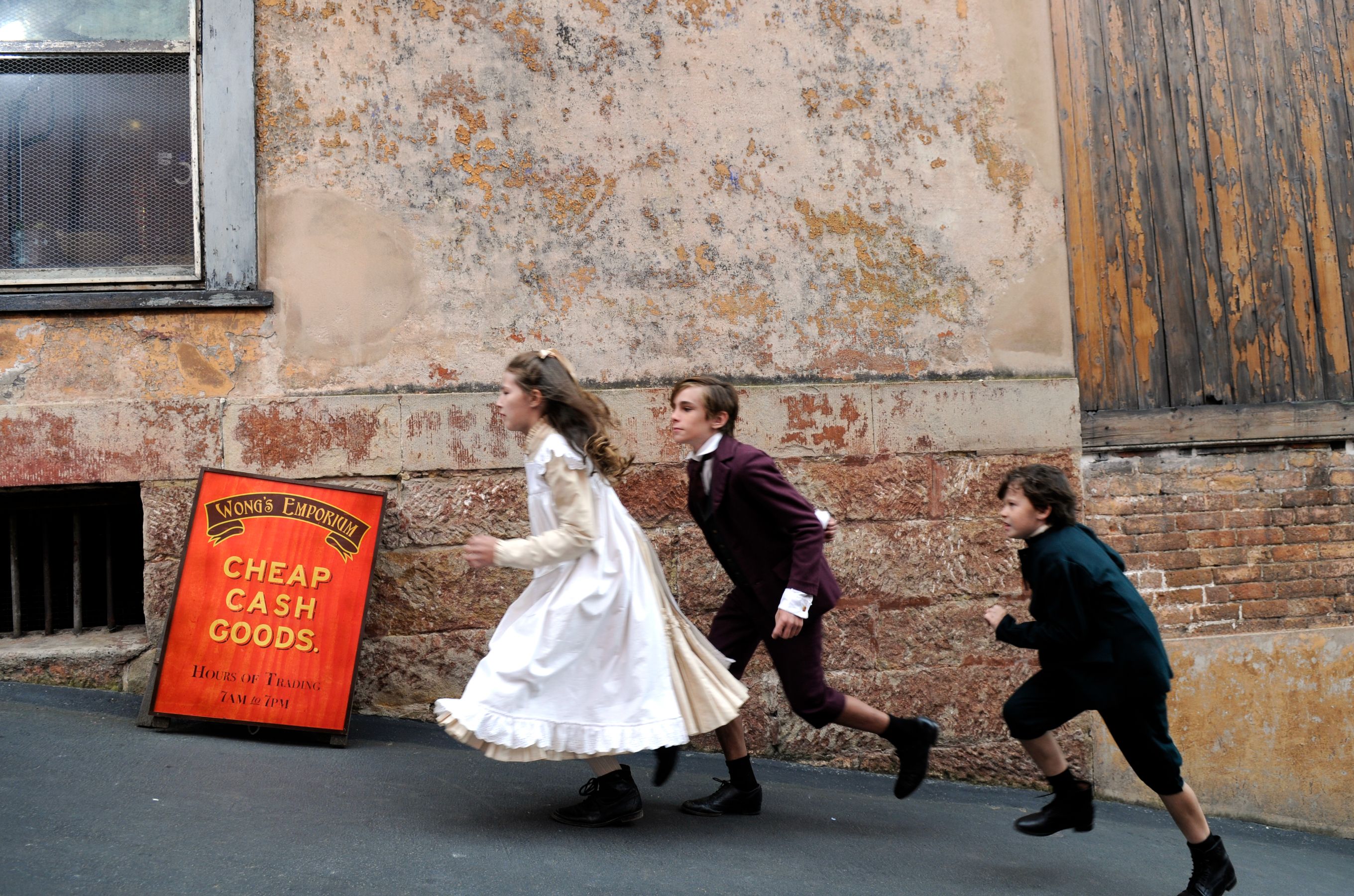 Three children are running along a weathered wall in a narrow alley. The girl, wearing a white dress with frills, leads the way, followed closely by two boys dressed in period clothing. A sign for "Wong's Emporium" advertising "Cheap Cash Goods" stands propped against the wall.