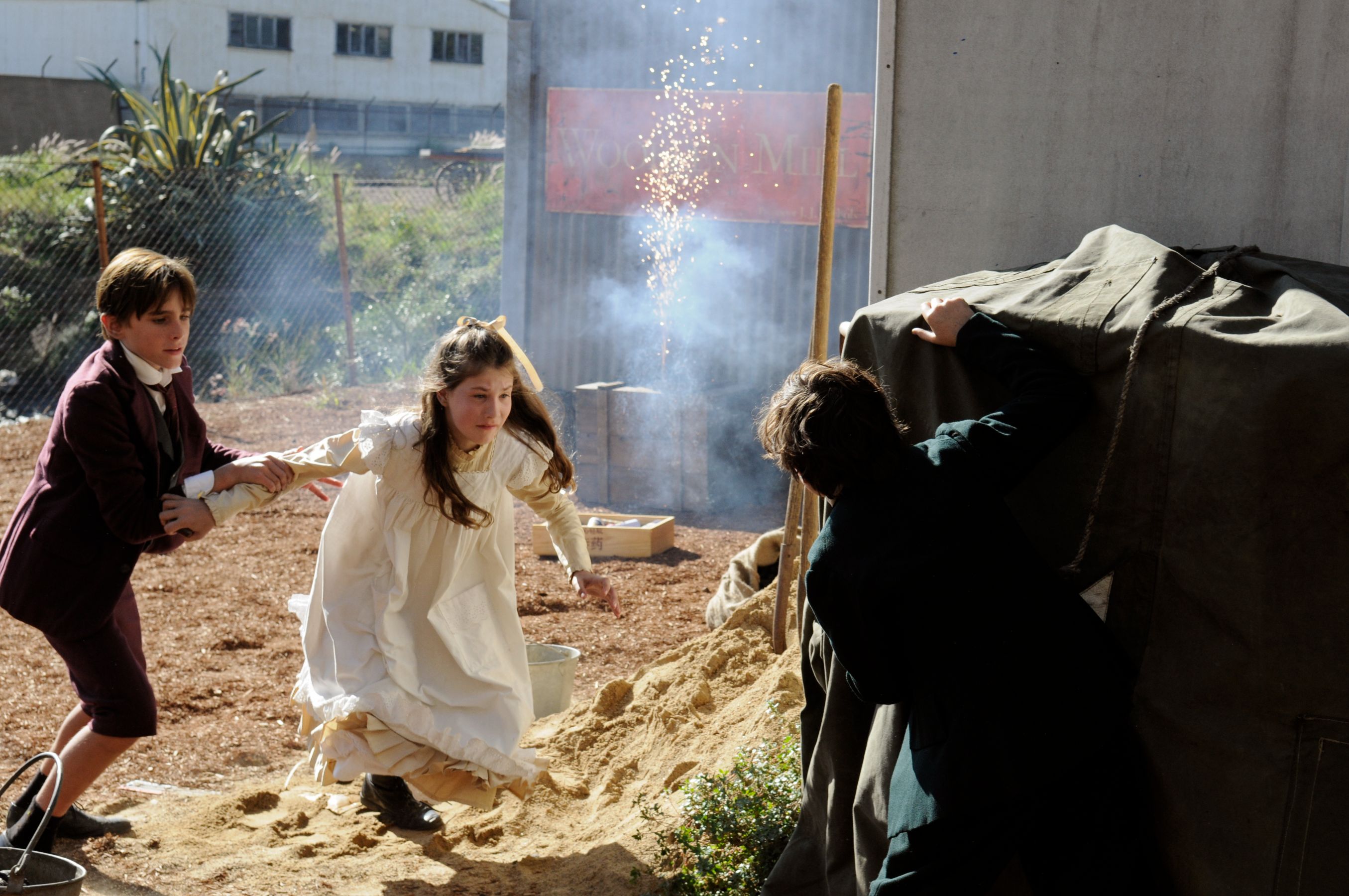 Two children, a boy in a dark suit and a girl in a white dress, appear to be fleeing from an unseen danger, with the boy pulling the girl along. In the background, smoke billows from a structure, and a flicker of sparks can be seen, suggesting an explosive event nearby.