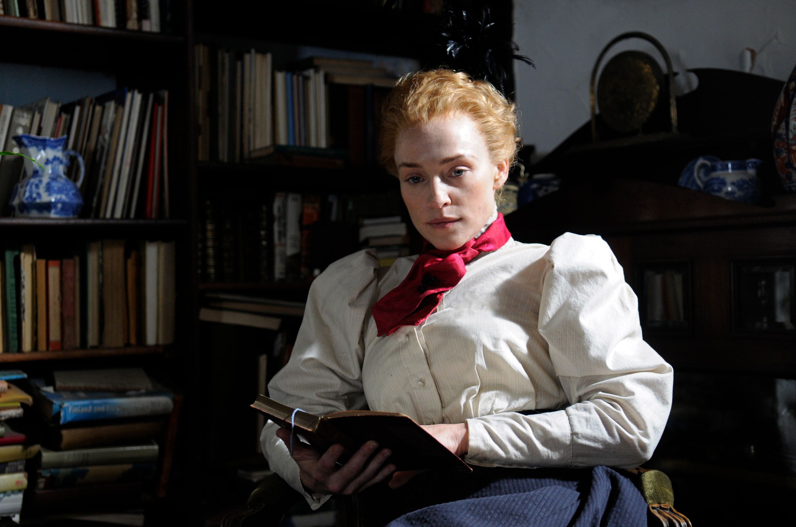 A woman with curly red hair sits in a dimly lit room, holding an open book in her lap. She wears a white blouse with puffed sleeves and a red necktie, her expression contemplative as she gazes downward. Behind her, shelves filled with books create a scholarly backdrop.