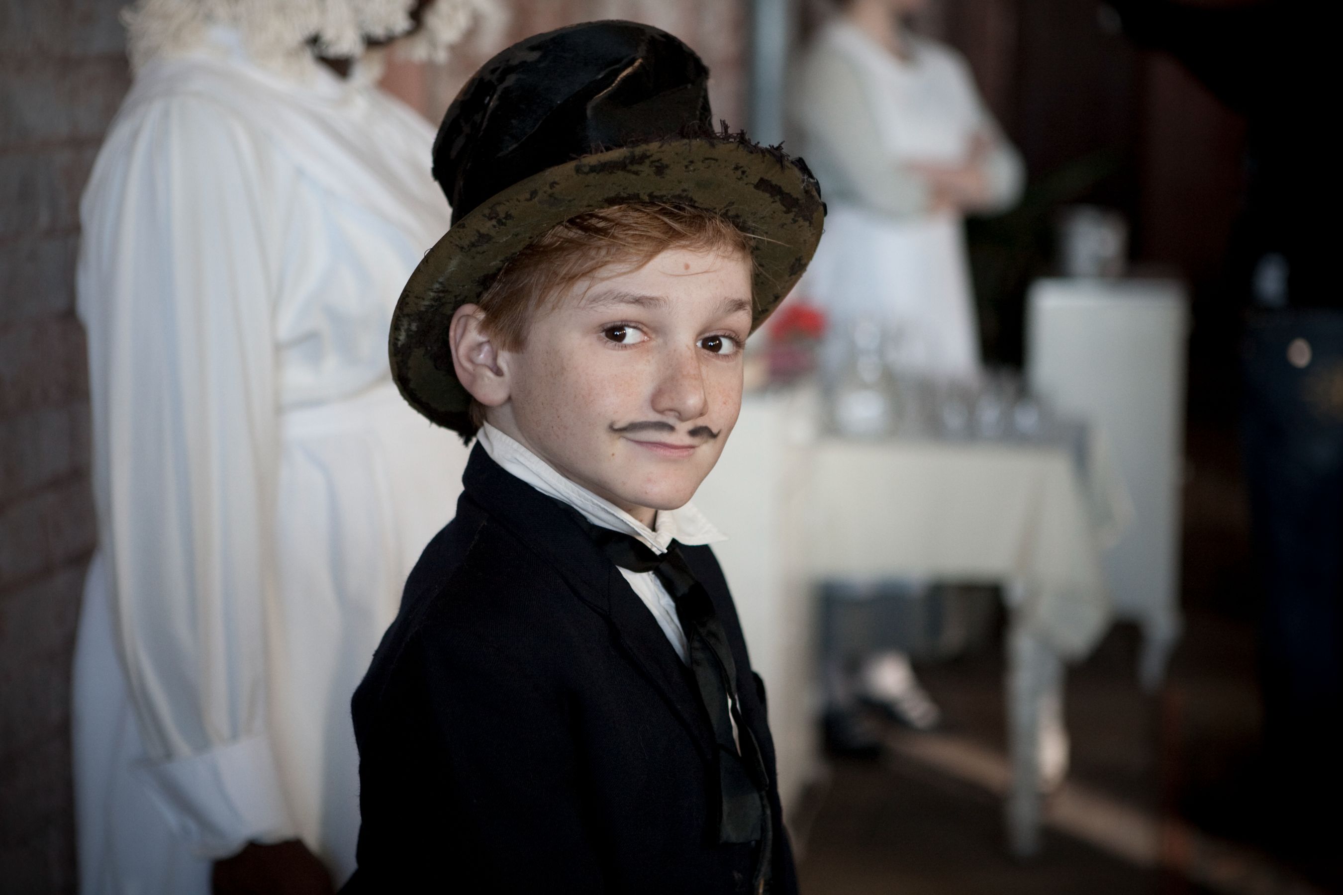 A young boy dressed in a formal black suit and top hat poses with a playful smile, sporting a small mustache. In the background, a character in a white dress stands, creating a contrasting setting.