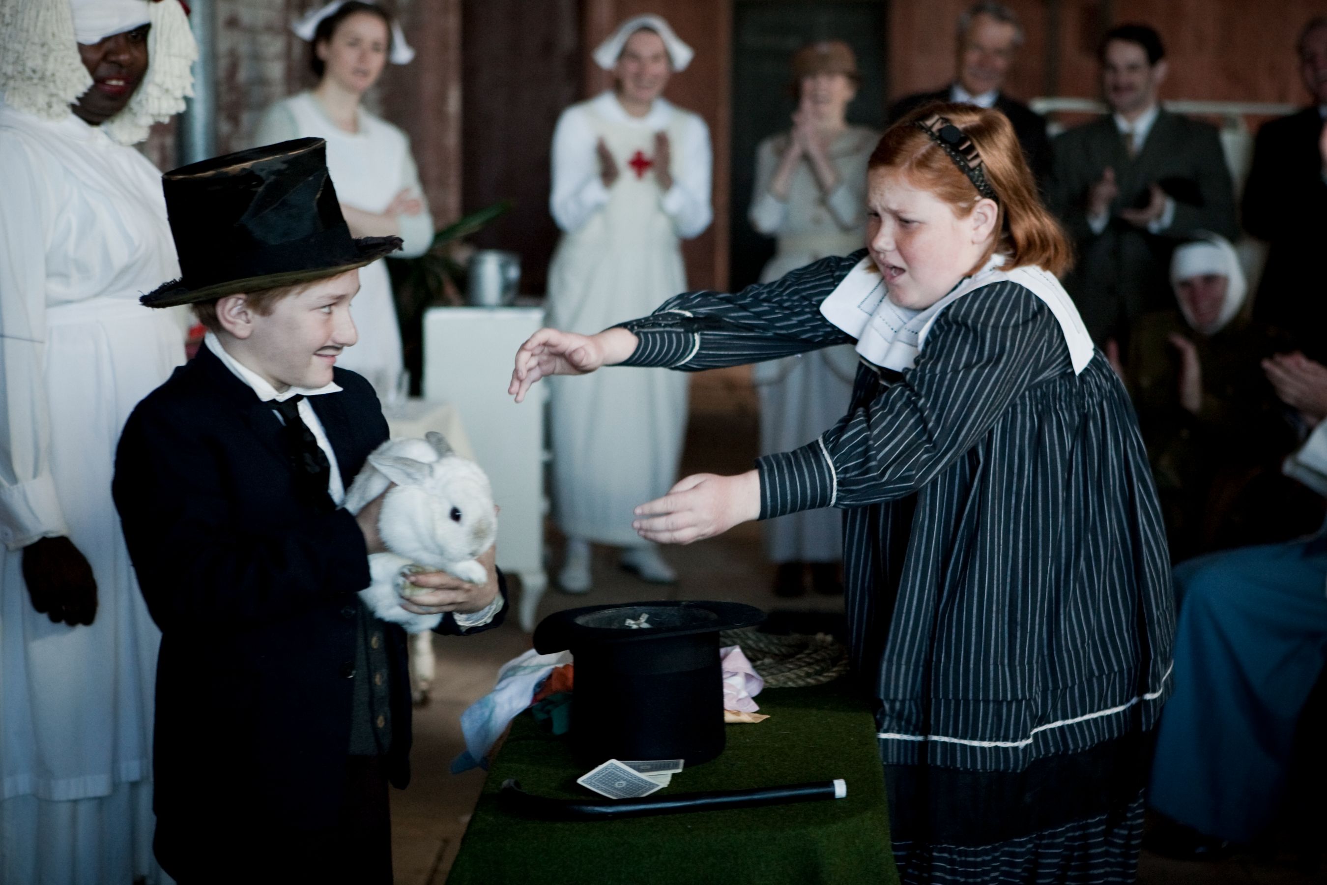 A young girl in a striped dress with a white collar appears surprised as she reaches for a rabbit held by a boy in a top hat and suit. In the background, an audience of dressed-up individuals watches with smiles and applause, creating a festive atmosphere.