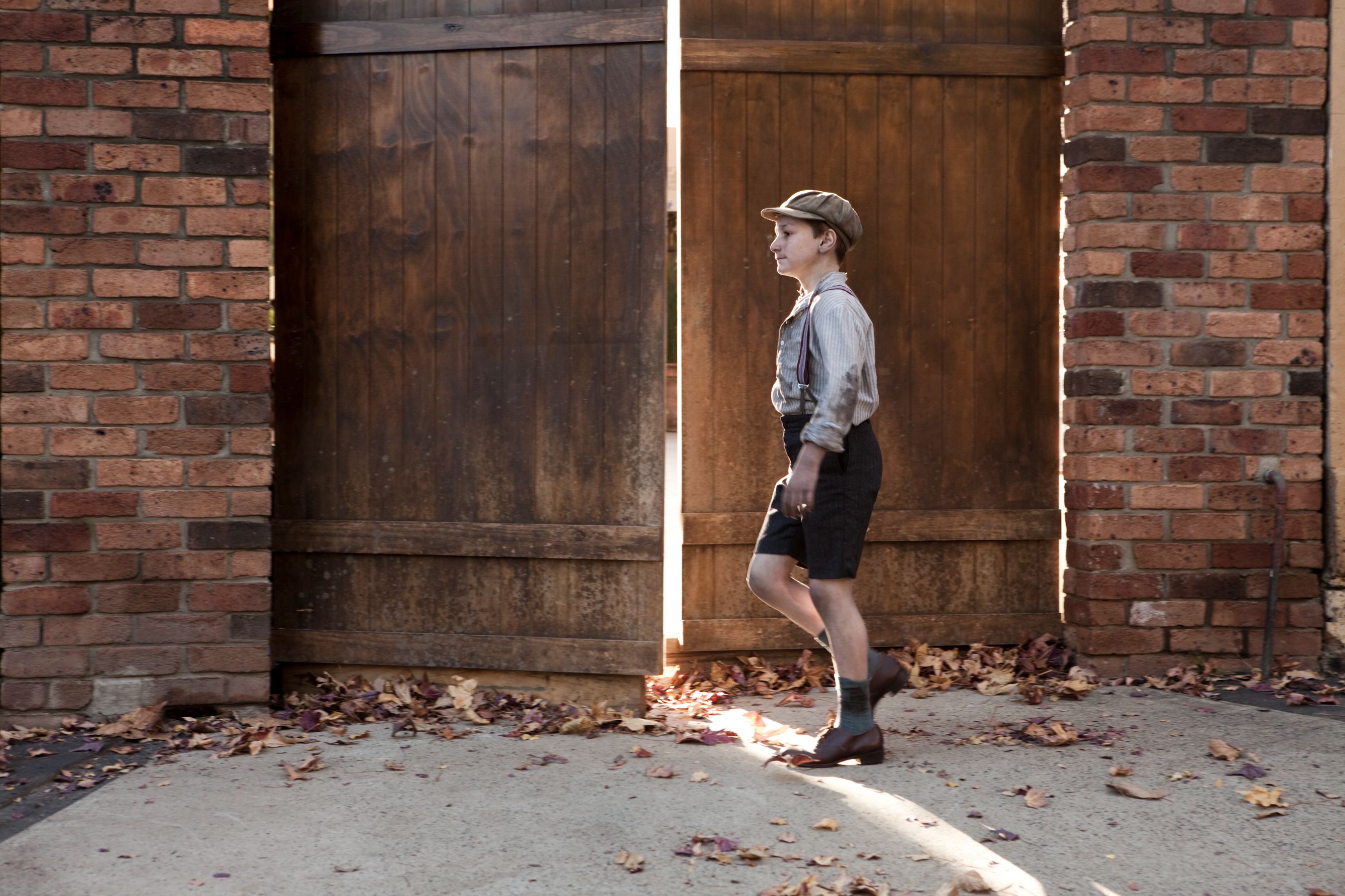 A person wearing a vintage outfit with suspenders, a button-up shirt, and a cap strolls along a path lined with fallen leaves, passing through large wooden gates flanked by brick walls. The scene is bathed in warm light, highlighting the nostalgic atmosphere.