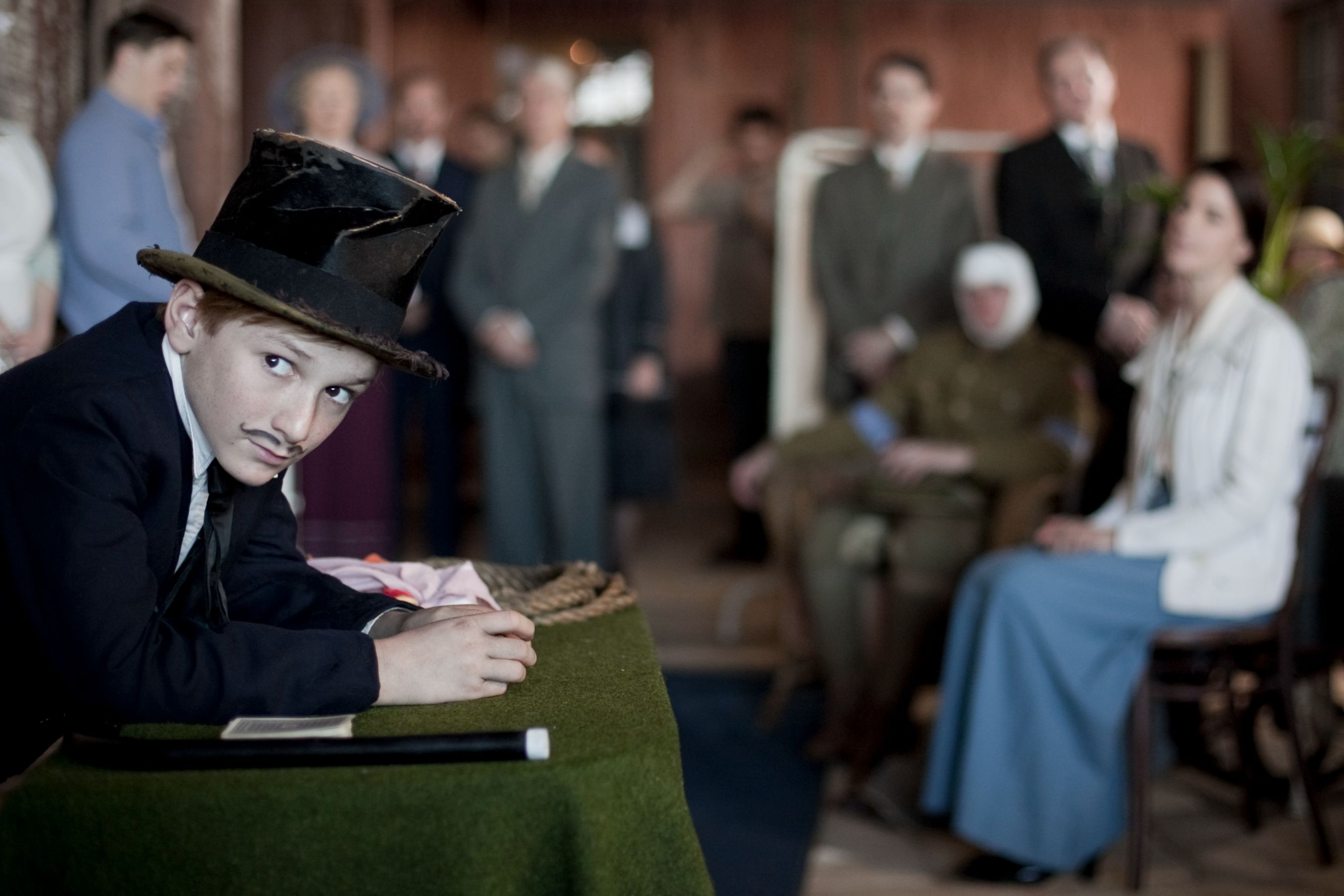 A young boy in a top hat with a thin mustache leans on a table covered in green fabric, gazing toward a group of well-dressed adults in the background. They observe him with a mix of curiosity and attention, while a woman sits nearby wearing a white blouse and blue skirt.