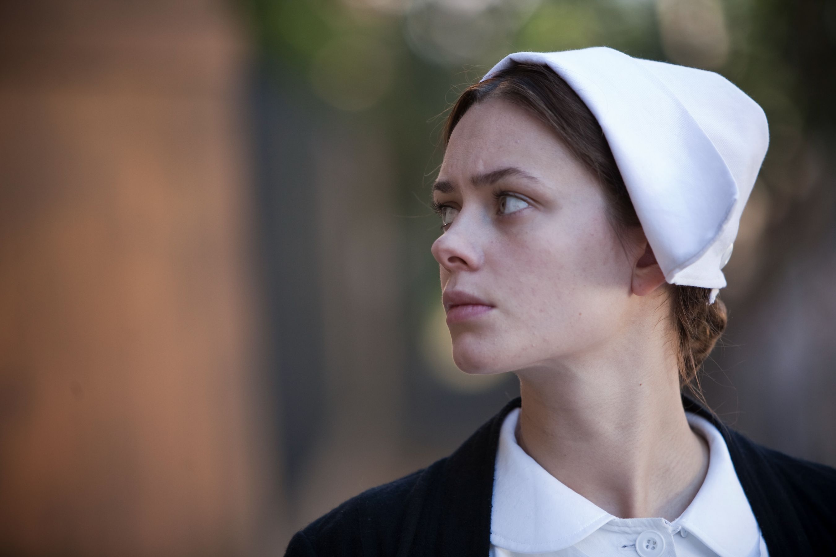 A woman with a serious expression wears a white bonnet and a black garment, looking off to the side. The background features blurred natural elements, suggesting an outdoor setting.