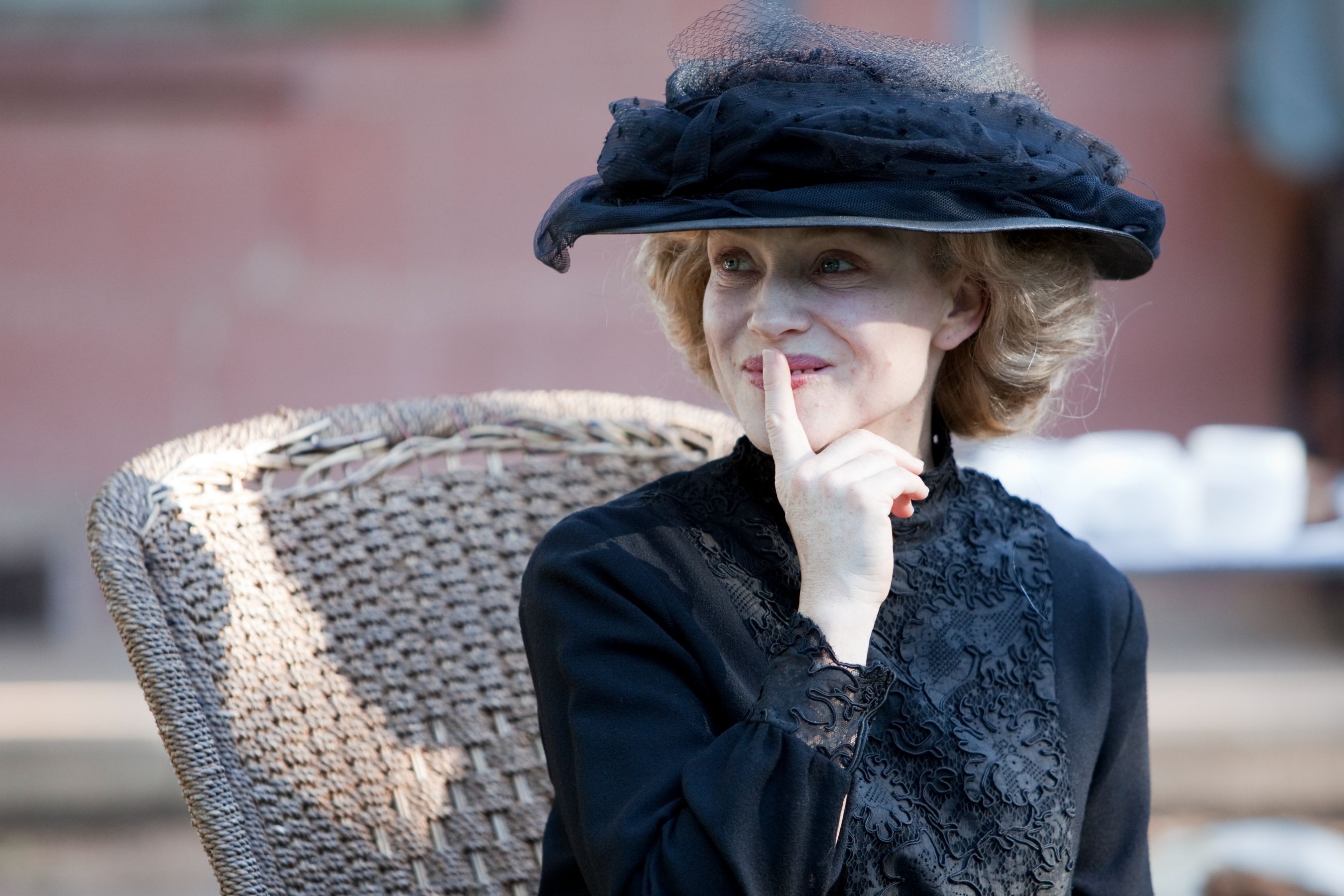 A woman sits in a woven chair, wearing a black hat with a veil and a black, intricately decorated dress. She holds her finger to her lips with a playful expression, suggesting secrecy or contemplation.