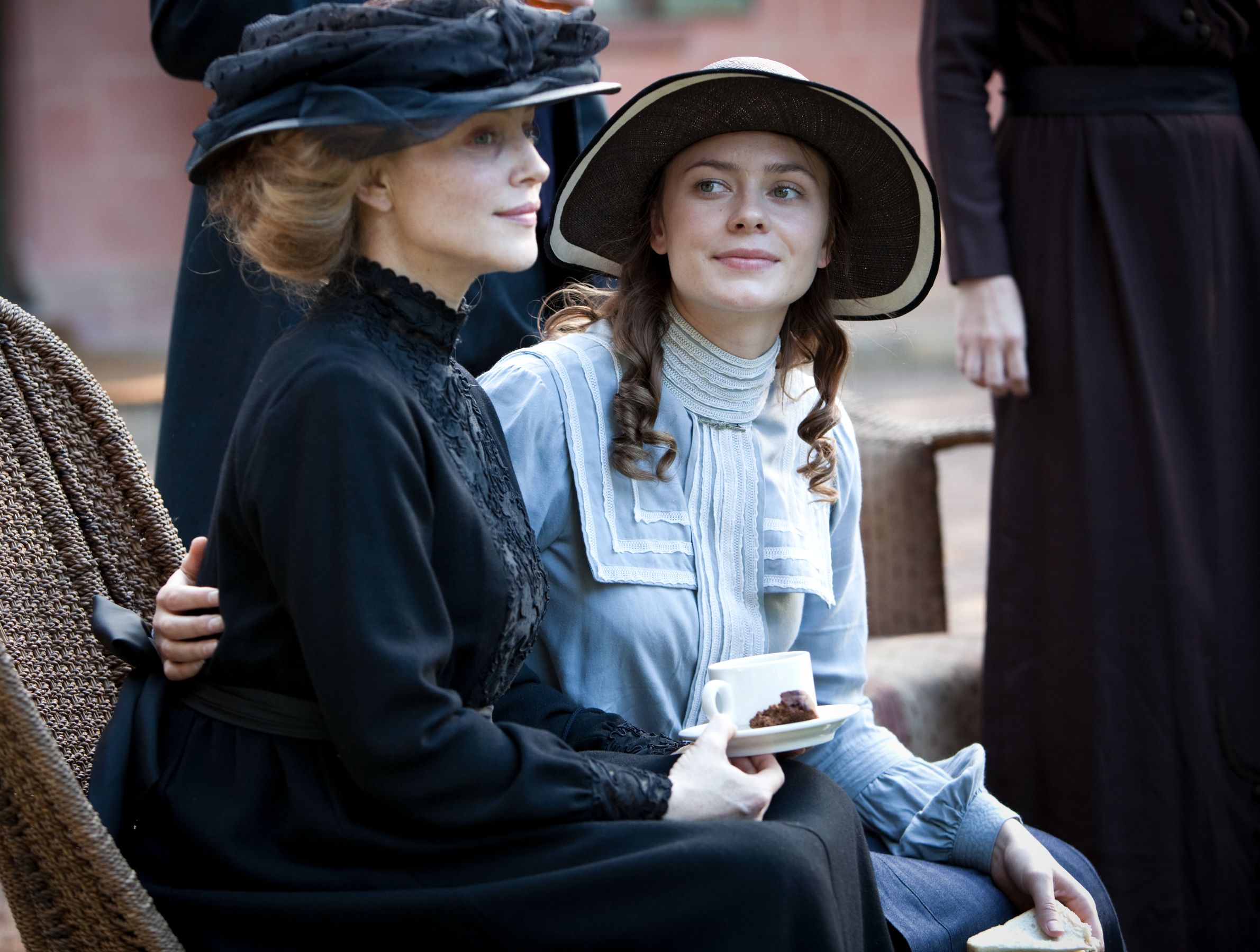 Two women are seated outdoors, both dressed in period clothing. One woman, wearing a black dress and a large hat, sits with a thoughtful expression, while the other, in a light blue blouse and an elegant hat, smiles warmly as she holds a cup and a dessert. A figure in a dark dress stands slightly behind them.