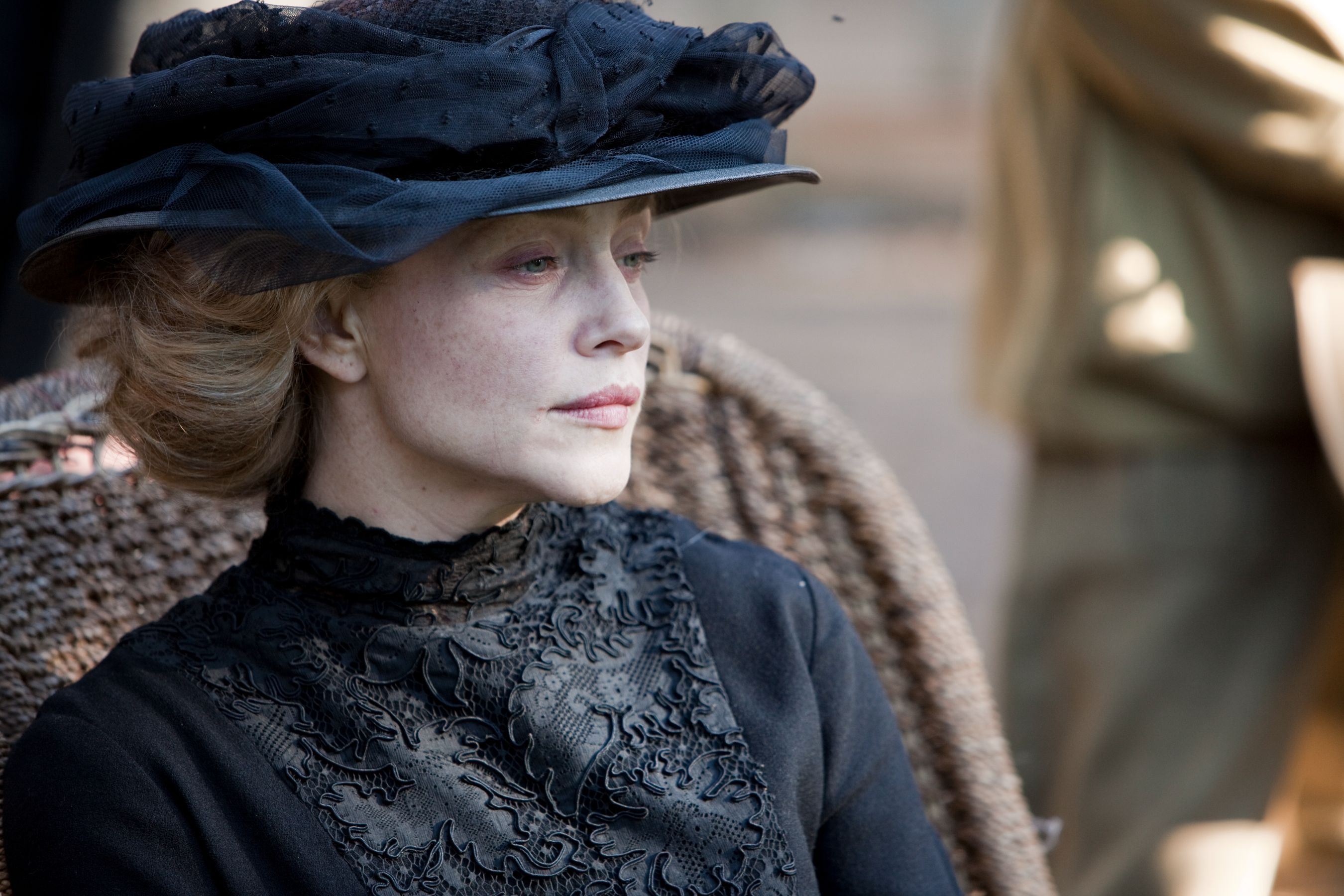 A woman with pale skin and delicate features gazes thoughtfully to the side, wearing a large black hat decorated with ribbons and a dark, intricately patterned dress. Her hair is styled in soft waves, contrasting with the somber colors of her attire against a blurred background.